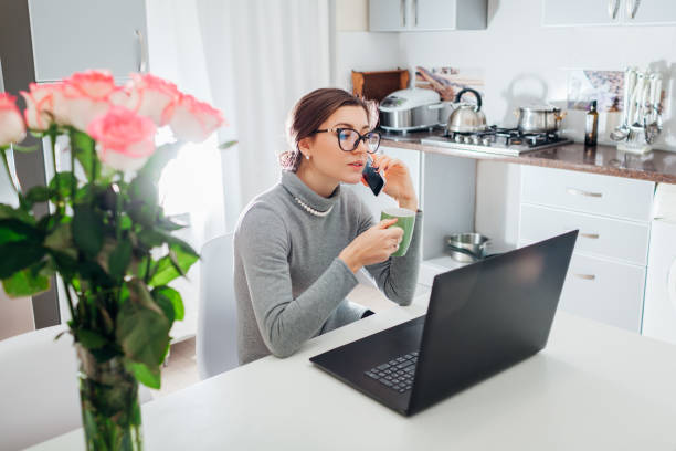 istock Woman talking on phone and drinking tea while working on laptop on modern kitchen. Freelancer relaxing 1077733562