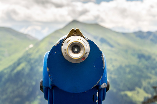 Public blue telescope arrayed against mountain range in Alps, Austria, cloudy summer day, travel wanderlust concept