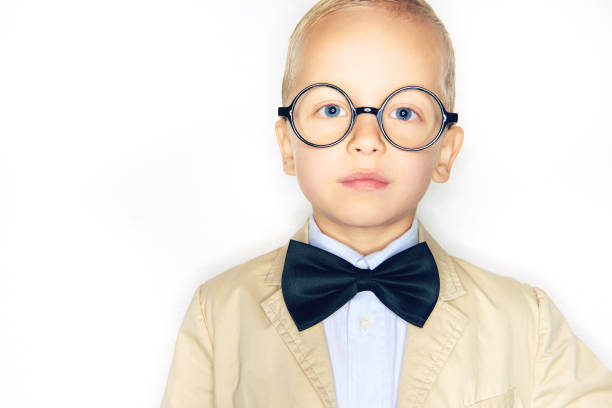 Adorable little blonde boy dressed like a professor wearing glasses, a suit and bowtie standing confidently against a white background