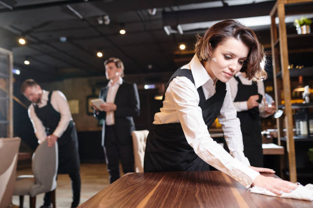 Busy waitress preparing restaurant for open and making cleanup Serious busy young waitress and her colleagues preparing restaurant for open and making cleanup: she wiping wooden table with cloth restaurant cleaning stock pictures, royalty-free photos & images