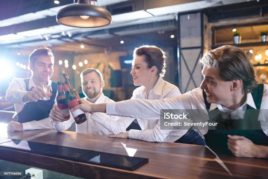 Excited restaurant colleagues clinking beer bottles at bar count Happy excited young restaurant colleagues reaching hands with beers to toast for good job, they sitting at at bar counter Adult Stock Photo