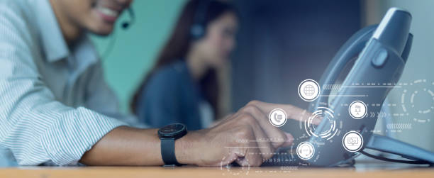 A close-up of a person wearing a watch and headset, smiling as they press buttons on a desk phone, with a blurred figure in the background and digital icons overlaying the image that suggest communication technology.