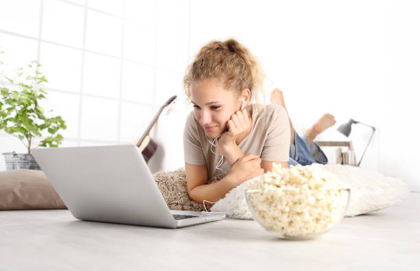 hermosa joven sonriente ver una película en la computadora comiendo palomitas acostado en el piso de madera de sala de estar en casa cómoda - lying down women laptop freedom fotografías e imágenes de stock