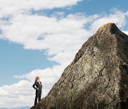 A businesswoman places her hands on her hips as she looks up at a tall mountain peak that stands in front of her.