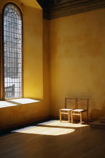 A long, narrow leaded glass window illuminates two chairs and a golden hued wall.