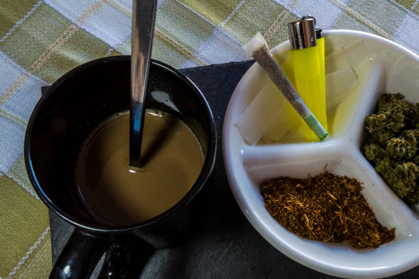 istock Materials for rolling joints: high quality marijuana buds, chopped tobacco, lighter and paper. Plate accompanied by a joint and a cup of coffee with milk. 1077824244