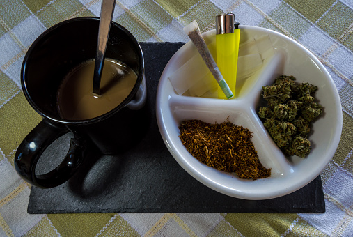 Materials for rolling joints: high quality marijuana buds, chopped tobacco, lighter and paper. Plate accompanied by a joint and a cup of coffee with milk.