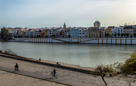 Views of guadalquivir river walk and buildings with people resting and riding bikes. Cloudy day of winter, Seville, Spain, February 2018.