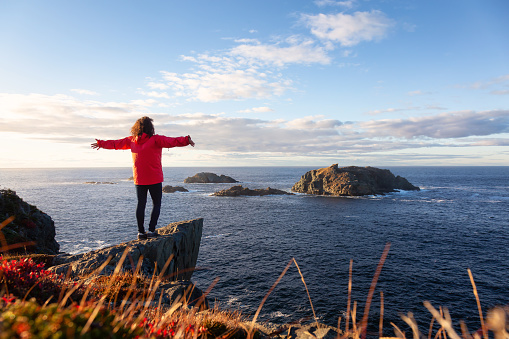Woman in red jacket is standing at the edge of a cliff with open arms and enjoying the beautiful ocean scenery. Taken in Crow Head, North Twillingate Island, Newfoundland and Labrador, Canada.