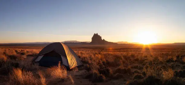 Striking panoramic landscape view of a tent in the dry desert with a mountain peak in the background during a vibrant sunset. Taken at Shiprock, New Mexico, United States.