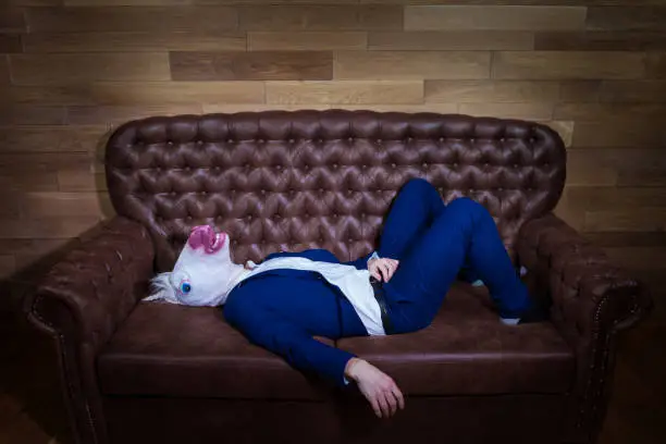 Funny unicorn in elegant suit lies on sofa. Unusual man at home. Freaky guy in comical mask on background of wooden wall.