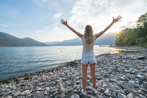 Cheerful young woman embracing nature at sunset; female standing on lakeshore with mountain background- Switzerland