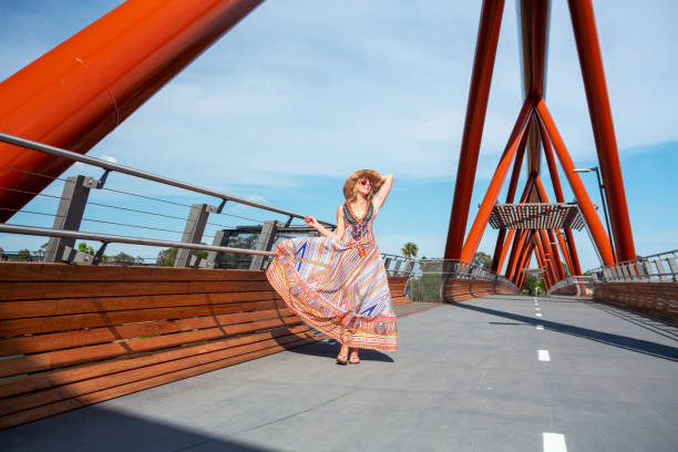 Woman on striking bridge wearing long maxi dress swaying Female wearing a  long summer sway dress of vibrant colours and straw hat, walking across the new bridge Yandhai Nepean Crossing Maxi Dress stock pictures, royalty-free photos & images