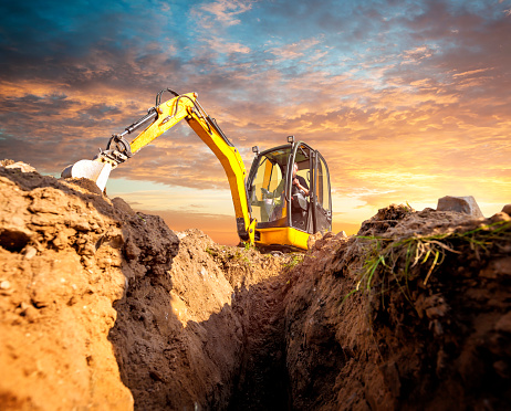 Mini Excavator. Yellow mini excavator on the construction site. Excavator operator chooses the ground for the foundation. Mini digger digs down in the background of the sunset.