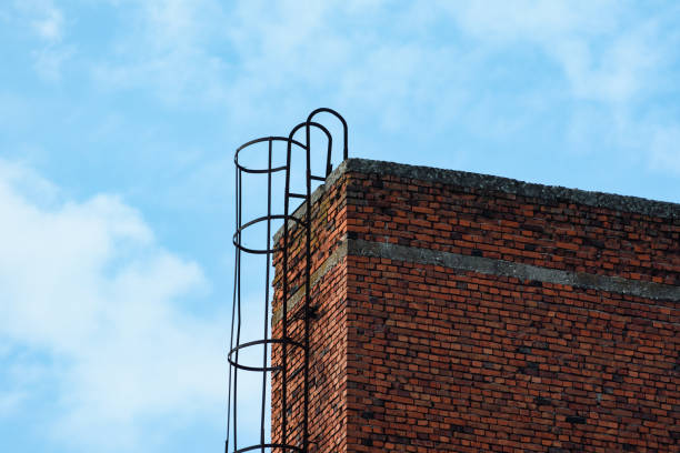 A low angle view of red brick wall with a rusty metal ladder against the cloudy blue sky.