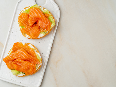 Two open sandwiches with salmon, cream cheese, cucumber slices on white marble table, copy space, top view