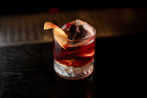 View from above glass of a Boulevardier cocktail with big ice cube and orange zest on the steel wooden bar counter on the blurred background