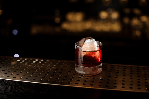 Glass of a Boulevardier cocktail with big ice cube on the steel wooden bar counter on the blurred background