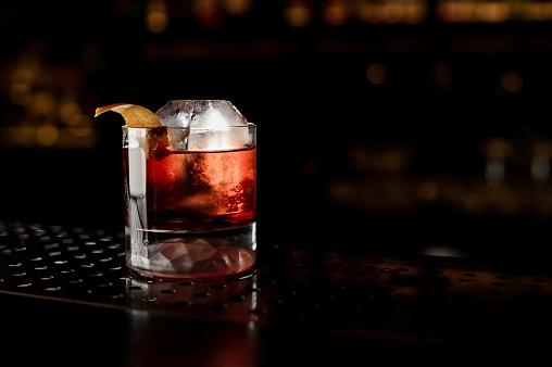 Glass of a Boulevardier cocktail with big ice cube and orange zest on the steel bar counter on the dark blurred background