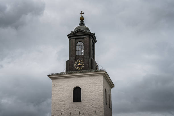 Top view of a very old church tower made of stone with a golden clock dial, located on the Swedish countryside. Against a dark gray cloudy sky from an incoming storm