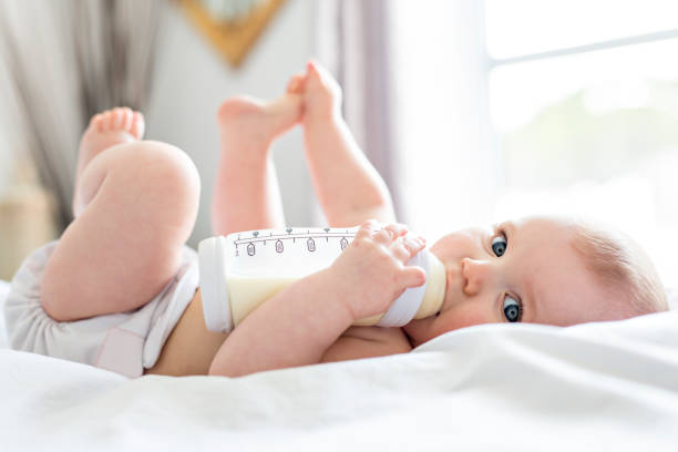 bonita niña bebe agua de botella acostado en cama. pañal jugar de niño en la sala vivero. - biberón fotografías e imágenes de stock