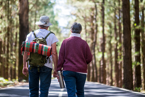Couple of old aged senior traveler walk in the middle of a road with forest around and backpack with blanket on the back. Love forever partnership and natural free lifestyle concept with no age limits concept