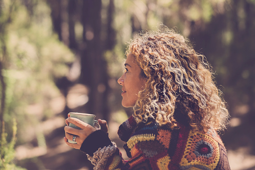 Beautiful woman in autumn forest enjoying the feeling with the nature with a warm sweater - lady sits with trees in background in a forest and holds a cup with a hot drink in her hands. Girl travel and wanderlust millennial concept
