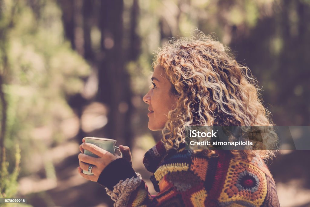 Beautiful woman in autumn forest enjoying the feeling with the nature with a warm sweater - lady sits with trees in background in a forest and holds a cup with a hot drink in her hands. Girl travel and wanderlust millennial concept Women Stock Photo