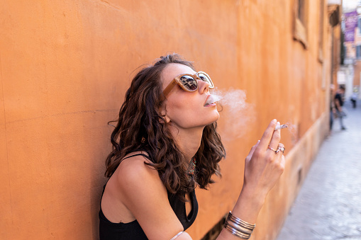 Side view of young woman with sunglasses smoking cigarette on street.