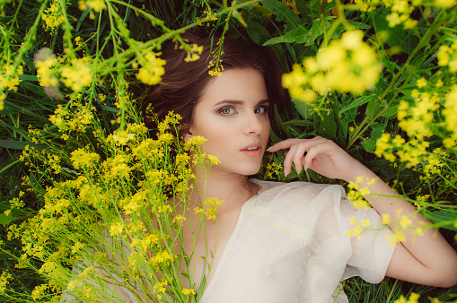 Portrait of young beautiful girl laying in field of yellow flowers with fingers near mouth and bouquet in hand looking to the camera