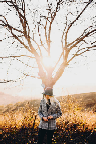 istock Beautiful young lady in elegant hat and jacket keeping eyes closed while standing near tree on sunny day in autumn nature.Stylish woman standing near naked tree 1078126684