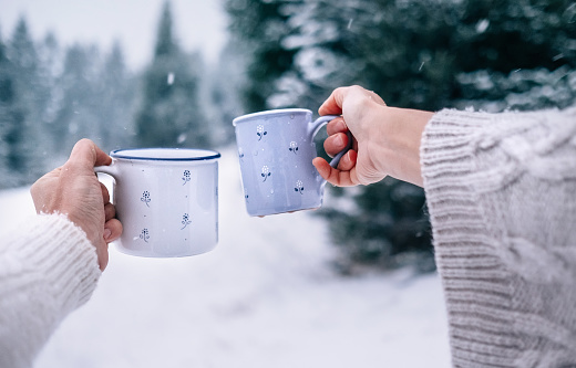 Man and woman hands in knitting mittens taking cups of hot drink. Sunny winter forest glade landscape on background. Winter forest concept image.