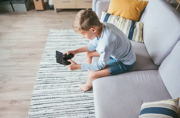 Preteen boy losted in computer game. Sits at home on coze sofa, uses gamepad with smartphone. Top view.