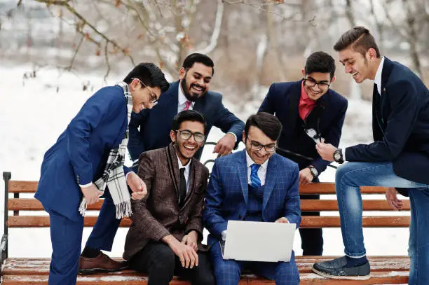 Group of six indian businessman in suits posed outdoor in winter day at Europe, looking on laptop and laughing.