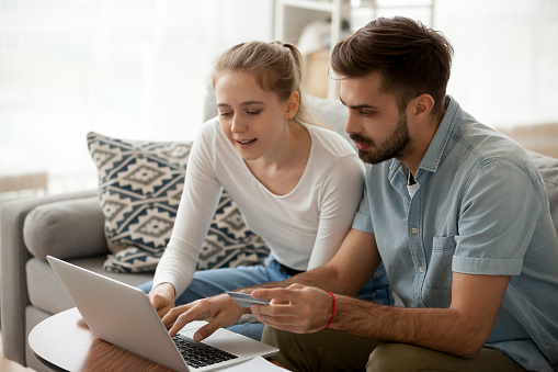 Millennial couple sit on couch at home shopping online together, man and woman enter credit card details making purchase in internet, husband and wife using computer contacting bank customer service