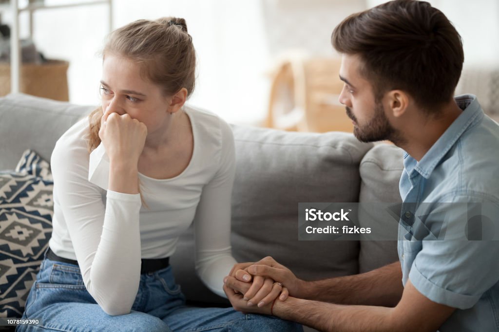 Caring husband hold wife hand making peace after fight Loving young husband hold crying wife hand showing empathy and support, millennial couple sit on couch at home reconcile after fight, caring man making peace with beloved woman. Relationships concept Couple - Relationship Stock Photo