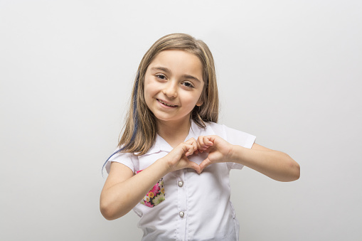 Close up portrait of little girl with beaming smile showing love symbol with fingers, looking at camera standing isolated on white background