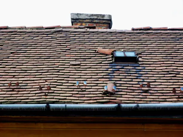 damaged sloped clay roof with decaying and breaking clay roof tiles, brick chimney and old galvanized steel gutter eavestrough