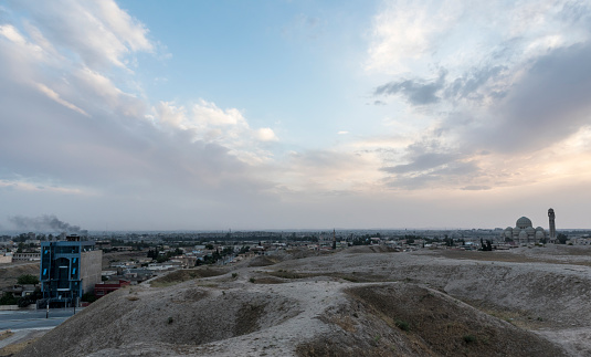 A view at dusk from atop the Nineveh ruins, looking toward the old city of Mosul, Iraq. In the center of this photo, far in the distance is the slightly left-leaning minaret of The Great Mosque of al-Nuri. On the left is smoke from the fighting with ISIS. On the right is the Grand Mosque of Mosul. (May 2017)
