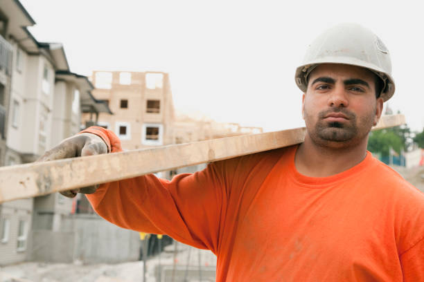 A skilled worker is measuring a wooden beam with a level tool, ensuring accuracy while building a frame structure. Safety gear and focused expression are evident.
