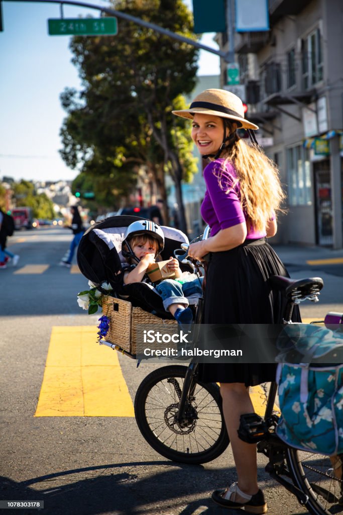 Mom Pushing Kid in Cargo Bike Smiling Back at Camera A young, stylishly dressed mother riding a "cycle truck" style cargo bike through the streets of San Francisco with a toddler strapped into a child seat over the front wheel. 2-3 Years Stock Photo