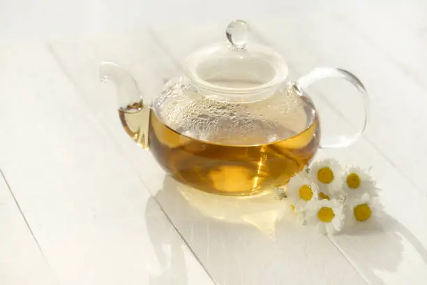 Chamomile tea in a transparent glass teapots and white daisy flowers on a white wooden background.Organic teas for health