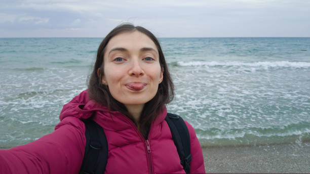 A girl in a warm jacket makes a selfie on the beach in windy stormy weather. Big wave. Girl showing tongue at the camera, boasting stock photo
