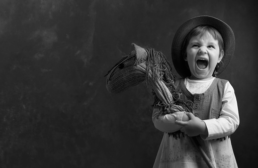 Screaming little girl with a homemade toy. Little girl dressed like a cowboy playing with a homemade horse. Expressive facial expressions. Copy space, black and white.