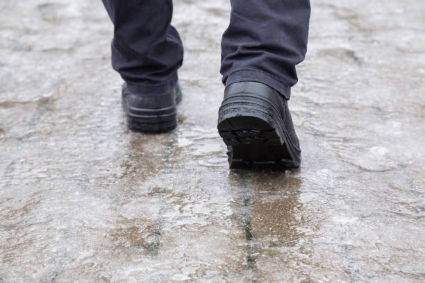 las piernas del joven en cuero negro botas caminando por la acera en húmedo, cálido día de invierno. pavimento cubierto de hielo resbaladizo. closeup. vista posterior. - road ice danger winter fotografías e imágenes de stock