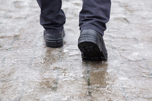 Young man's legs in black leather boots walking on sidewalk in wet, warm winter day. Pavement covered with slippery ice. Closeup. Back view.