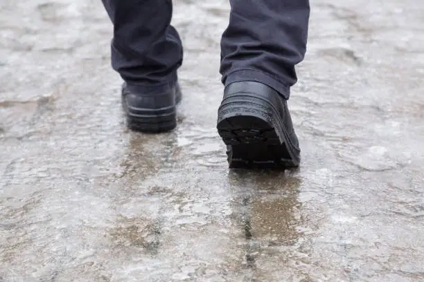 Young man's legs in black leather boots walking on sidewalk in wet, warm winter day. Pavement covered with slippery ice. Closeup. Back view.