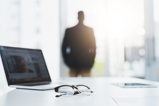 Closeup shot of a pair of spectacles and a laptop on a table in an office with a businessman in the background