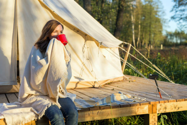 Young woman wraps blanket over herself while drinking coffee near canvas tent in the morning Young woman wraps blanket over herself while sitting and drinking coffee near canvas tent in the morning in the woods glamping photos stock pictures, royalty-free photos & images