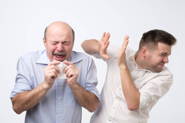 Poor old guy has terrible grippe. Studio shoot Sick bald caucasian man holding napkin or tissue, trying to cover mouth while sneezing with closed eyes. His coworker try to hide from microbes. Poor guy has terrible grippe sneezing stock pictures, royalty-free photos & images